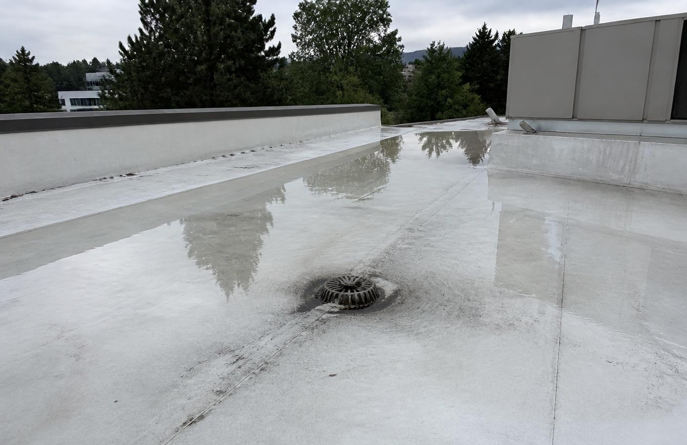 Ponding water around a roof drain on a white low-slope commercial membrane roof with a parapet wall and rooftop equipment nearby